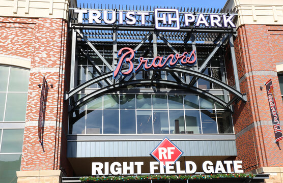 A vibrant exterior view of Truist Park, home of the Atlanta Braves, captured on a bright sunny day with dramatic clouds scattered across the blue sky. The massive stadium fa&ccedil;ade f