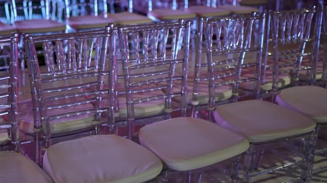 Rows of modern, clear chairs with cushions under elegant purple pink event lighting. Luxury banquet, wedding reception decor.