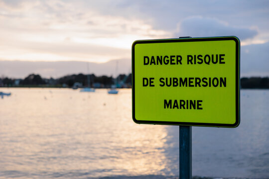 Bright yellow French coastal flooding warning sign reading &ldquo;danger risque de submersion marine&rdquo;, with blurred harbor and boats in soft evening light in background.