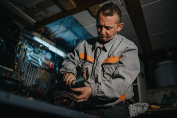 Naklejka premium A mechanic in a gray jumpsuit with orange accents concentrates on repairing a mechanical part in a busy workshop filled with tools, cables, and equipment, conveying skilled handiwork.