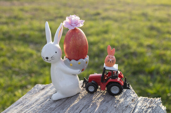 souvenir Easter bunny with painted red egg, toy model of tractor standing on tree bark in beautiful sunlight. commercial Easter greeting concept for farms and agribusinesses. Postcard. Copy space