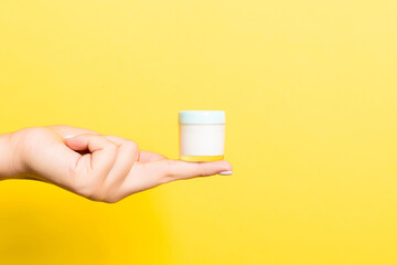 Female hand holding cream bottle of lotion isolated. Girl give jar cosmetic products on yellow background