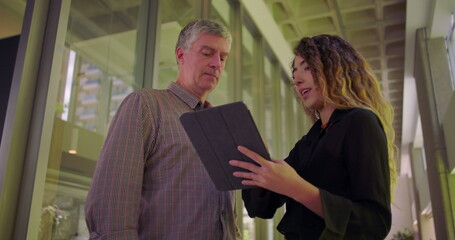 Man and woman in office hallway looking at tablet, discussing project with focused expressions, collaborative business meeting in shared workspace