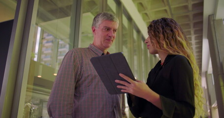 Man and woman in office hallway looking at tablet, discussing project with focused expressions, collaborative business meeting in shared workspace