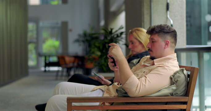 Man and woman sitting apart in modern indoor hallway using smartphones, both immersed in screens with neutral expressions, disconnected from each other