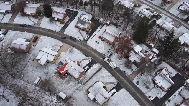 Drone shot looking down from aerial above garbage truck driving along neighborhood street and collecting trash, waste, and can junk by side of homes and house yards with claw during cold snowy winter
