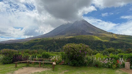 Scenic view of Mount Sinabung, North Sumatra, Indonesia. An active stratovolcano in the Karo Highlands on a cloudy day with a garden and an empty wooden bench in the foreground.