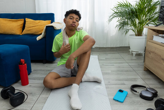Brown skinned teenager sitting on an exercise mat, sweating and looking tired while resting with a towel around his neck. A scene of home workout and recovery