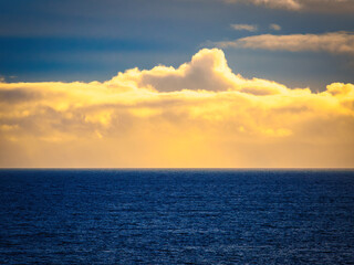 A vast, calm ocean stretches to the horizon. Golden sunlight shines through the clouds, creating a warm glow over the dark blue water during sunset.
