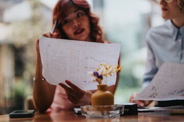 Two colleagues discuss charts and notes, holding printed sheets in a bright cafe setting. A warm, collaborative moment as they plan, share ideas, and evaluate data.