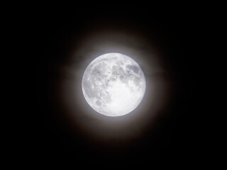 A bright full Moon dominates the dark night. It shows its craters and textures with a soft halo of light in a sky free of stars.