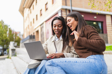 Two diverse women looking at a laptop outdoors, one smiling with surprise, representing friendship,...