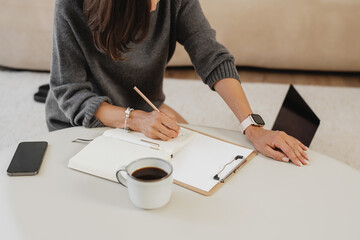 Woman writing in an open notebook at a low coffee table, cup of coffee nearby, clipboard and phone on the workspace.