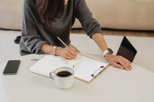 Woman writing in an open notebook at a low coffee table, cup of coffee nearby, clipboard and phone on the workspace.