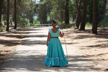 Fototapeta premium A beautiful young Spanish woman dressed in a traditional green flamenco dress with a flower in her hair walks among the pine trees at the pilgrimage, leaning on a stick. Photo taken from behind.