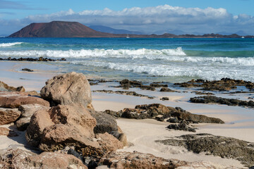 Lobos Island seen from the beach at Corralejo, Fuerteventura (Canary Islands). There are rocks and sand in the foreground. Mountains on the island of Lanzarote can be seen in the background.