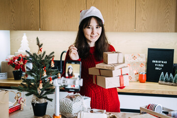 Young adult woman in red sweater reads handwritten Christmas shopping list on notebook next to small decorated tree. Social media gift guides, influencer unboxings, Christmas influencer marketing.