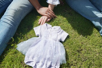 Fototapeta premium A couple sits on grass beside a small baby dress laid on the lawn, symbolizing pregnancy and parenthood. 