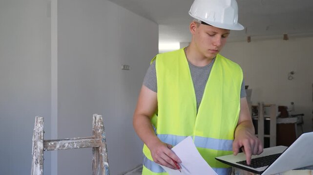 Teenage boy wearing yellow vest and hardhat using laptop and checking documentation in construction site. Young man having architect apprenticeship during part-time work.