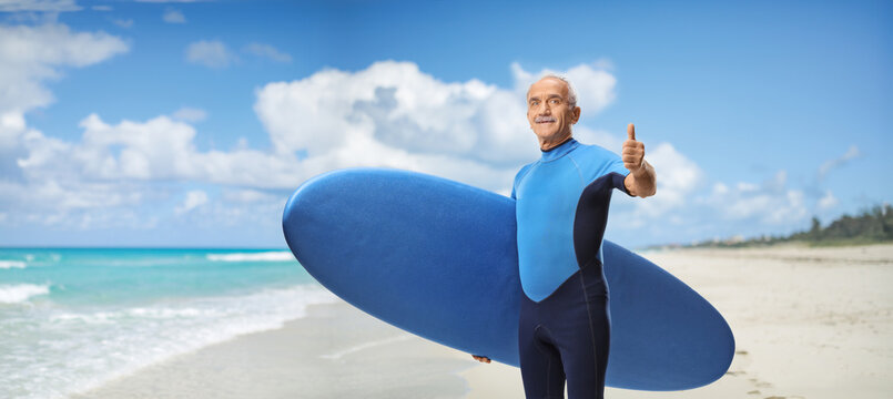 Elderly man in a wetsuit on a beach carrying a surfboard