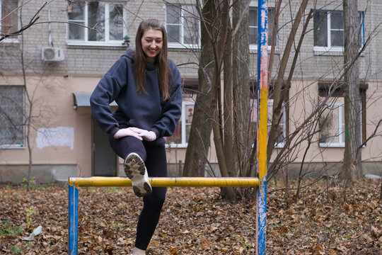 An outdoor workout area features a young woman stepping onto yellow parallel bars. One leg is lifted over the bar, and both hands rest firmly on the metal for balance.