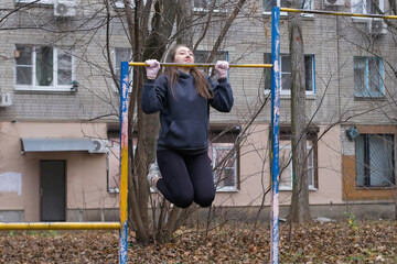 Naklejka premium European woman holds chin up position on pull up bar in city park, focusing on upper body and arm strength. Outdoor calisthenics workout, autumn setting, physical training and self improvement.