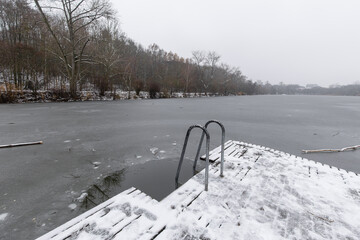 Snow covered wooden pier with metal ladder dipping into a frozen pond in winter. Horejsi rybnik in prague 9