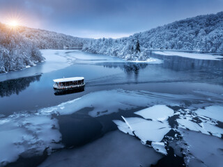 Naklejka premium Aerial drone view of boat in frozen lake at sunset. Winter fairytale in Plitvice Lakes, Croatia. Top view of boat, reflections in clear blue water, floating ice, snowy forest. White snow-covered trees