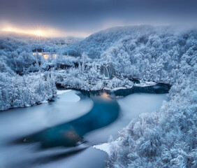 Naklejka premium Aerial drone view of frozen lakes and waterfalls surrounded by snowy forest at sunrise in Plitvice Lakes National Park, Croatia. Winter landscape with icy cascades and scenic European nature. Top view