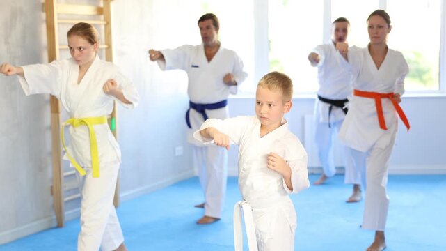 Boy with his family in kimonos and colored belts practicing karate with punches during group martial arts class in gym, accompanied by trainer
