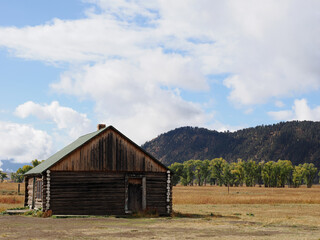 Obraz premium Rustic log pioneer cabin on wide prairie landscape with forested hill backdrop