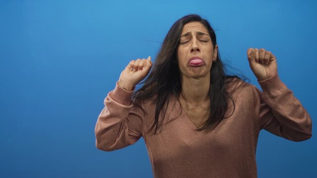 Woman wearing brown vneck sweater making a grimacing tongue out face with clenched fists and closed eyes in blue studio; disgust.