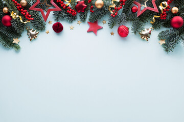 Fototapeta premium Festive winter frame with pine branches, red berries and shiny golden decorations on blue table top view. Christmas background in flat lay style.