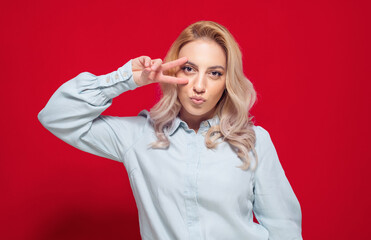 Pretty girl shows peace sing with fingers and send kiss to you, isolated on red background. Joyous lovely woman kissing