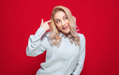 Funny woman makes finger gun gesture in temple, pretends committing suicide, isolated on red background. Silly girl sticking out tongue