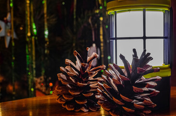 Camping lantern illuminating pine cones near Christmas tree lights