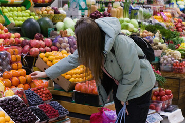 At a busy produce market, a young woman in a light green jacket reaches out to pick up fruit from a colorful selection. The scene is lively with a variety of fruits around her.