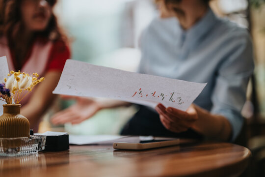 Two colleagues review a printed report full of graphs at a wooden table in a cafe. A smartphone rests nearby as they discuss data and planning in a relaxed, collaborative mood.