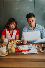 Two professionals sit at a wooden desk, examining papers and talking through notes as they collaborate on a project in a busy office setting.