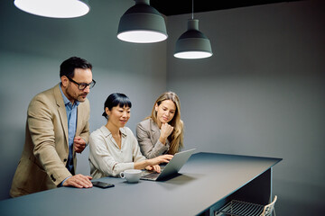 Three diverse business colleagues working together on a laptop in a contemporary office setting, discussing project ideas