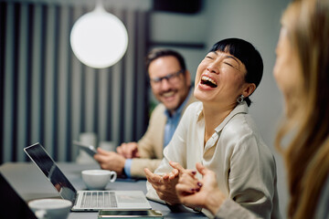 Asian businesswoman laughing while enjoying a business meeting with diverse colleagues in a modern office workplace