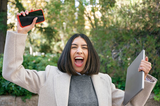 Happy woman celebrating success holding smartphone and laptop outdoors - Powered by Adobe
