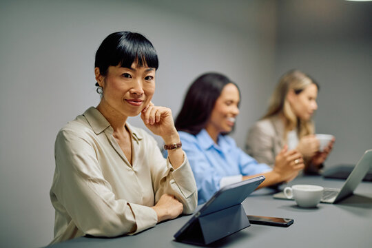 Asian businesswoman smiling at camera, sitting at table with tablet during a corporate meeting with diverse colleagues