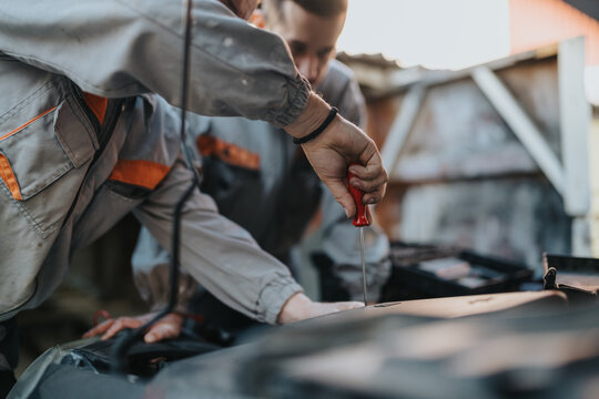 A mechanic in work wear leans over a car hood, using a red-handled screwdriver to tighten or inspect components. The scene captures hands-on automotive repair in an outdoor workshop. - Powered by Adobe
