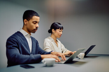 Two focused diverse business colleagues collaborating on laptops, managing projects and achieving shared company goals
