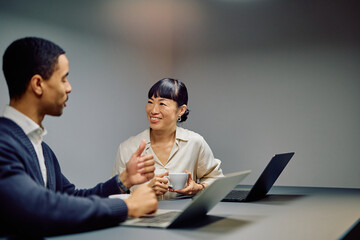 Multiracial business colleagues meeting, smiling, and discussing work while using laptops on a desk in a modern office