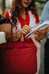 A group of professionals gather outside, discussing papers and sharing notes. The red bag and folders emphasize a collaborative, dynamic work moment.