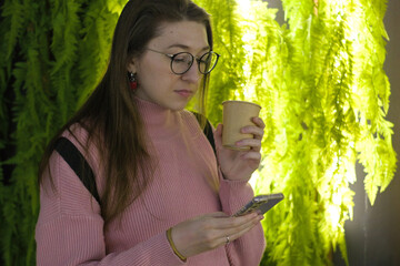 A young woman with glasses in a pink sweater stands by a green wall, holding a cup. She looks at her phone, appearing thoughtful and focused on the screen.