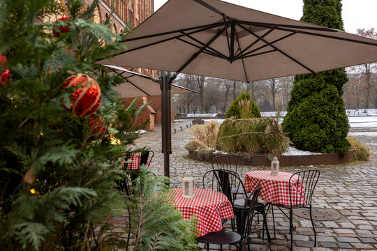 Outdoor cafe with red checkered tablecloths, metal chairs and umbrellas near decorated spruce tree with ornaments. - Powered by Adobe