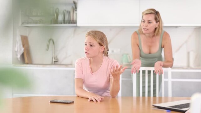 Saddened offended teen girl listening to disapproving words of mother standing in home kitchen. Concept of tension and disagreement arising between parents and child